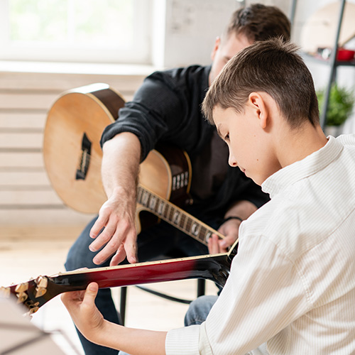 Gitarrenlehrer zeigt bei jugendlichem Schüler auf die Saiten seiner Gitarre