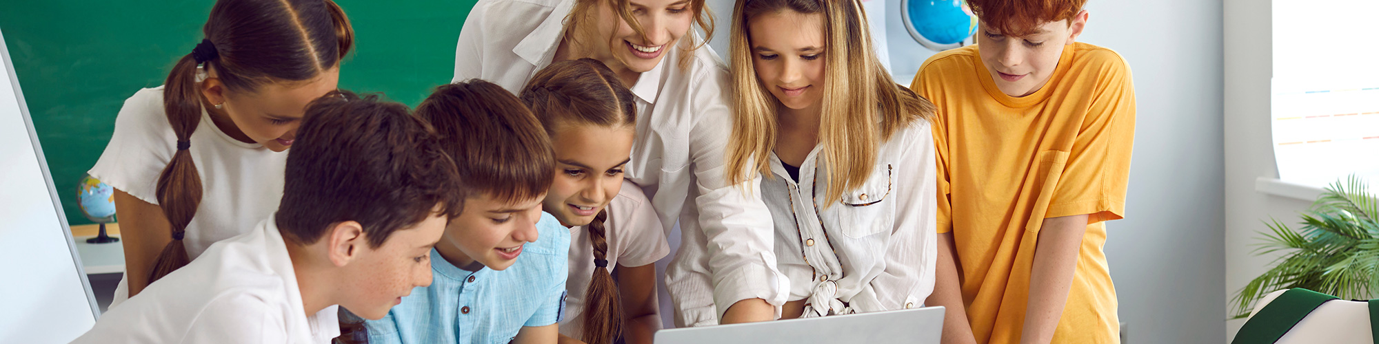 Sechs Kinder schauen vor einer Tafel mit der Lehrerin auf einen Laptop